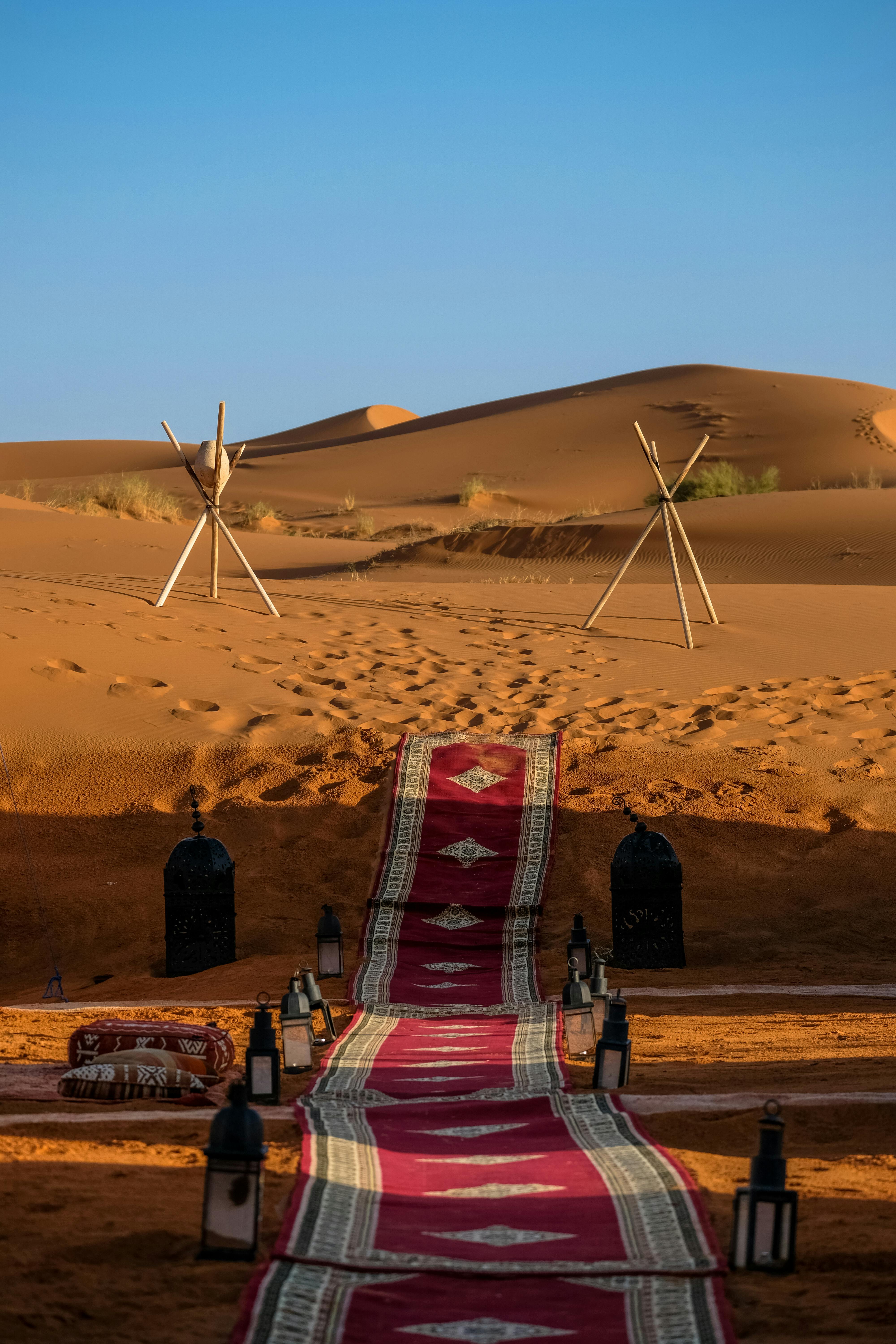 Desert setting with a red patterned rug, bottles, and two metal stands on sand dunes.
